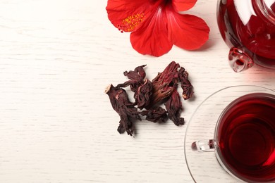Photo of Delicious hibiscus tea in glass cup, teapot, dry roselle sepals and flower on light wooden table, flat lay. Space for text