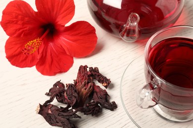 Photo of Delicious hibiscus tea in glass cup, teapot, dry roselle sepals and flower on light wooden table, closeup