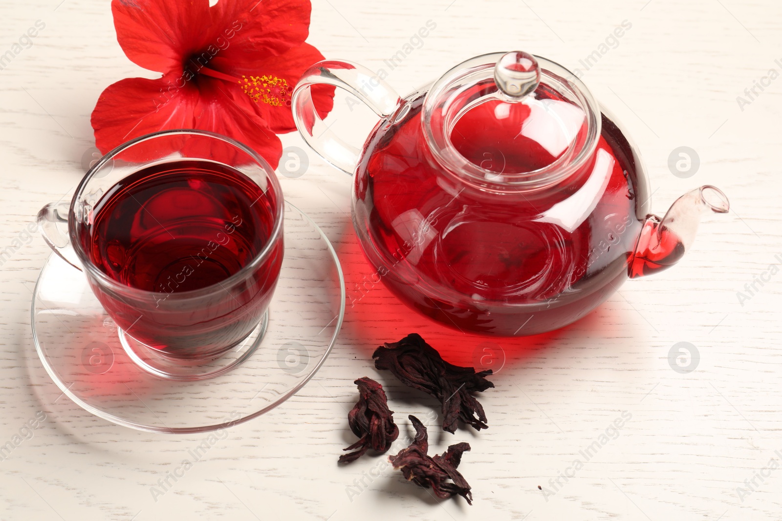 Delicious hibiscus tea in glass cup, teapot, dry roselle sepals and flower on light wooden table Photo of Delicious hibiscus tea in glass cup, teapot, dry roselle sepals and flower on light wooden table