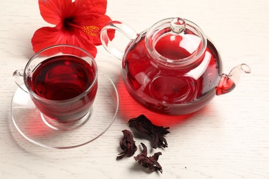 Photo of Delicious hibiscus tea in glass cup, teapot, dry roselle sepals and flower on light wooden table
