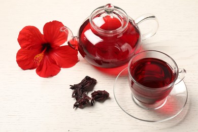 Photo of Delicious hibiscus tea in glass cup, teapot, dry roselle sepals and flower on light wooden table