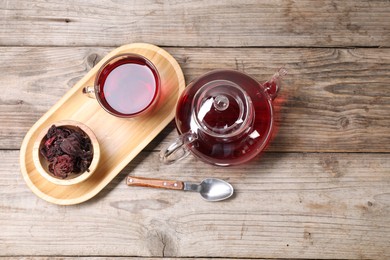 Photo of Delicious hibiscus tea in glass cup, teapot, dry roselle sepals and spoon on wooden table, flat lay. Space for text
