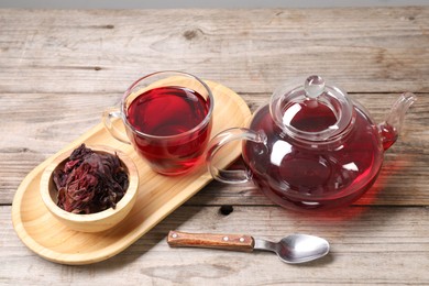 Photo of Delicious hibiscus tea in glass cup, teapot, dry roselle sepals and spoon on wooden table