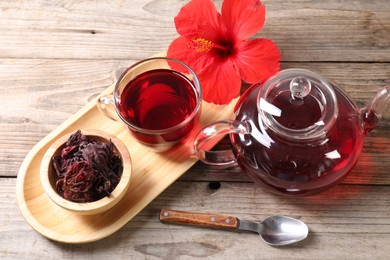 Photo of Delicious hibiscus tea in glass cup, teapot, dry roselle sepals and flower on wooden table