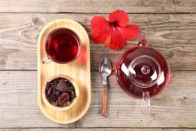 Photo of Delicious hibiscus tea in glass cup, teapot, dry roselle sepals and flower on wooden table, flat lay