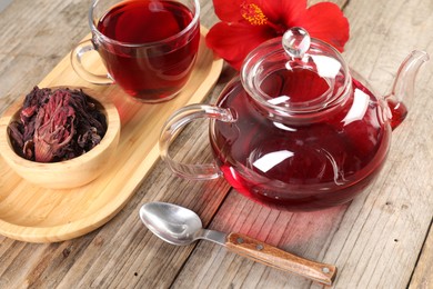 Photo of Delicious hibiscus tea in glass cup, teapot, dry roselle sepals and flower on wooden table, closeup