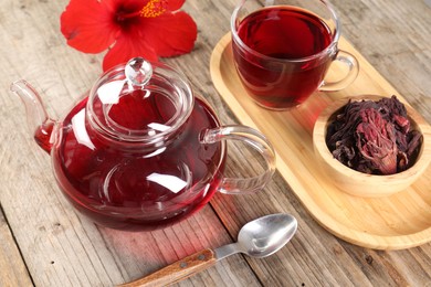 Photo of Delicious hibiscus tea in glass cup, teapot, dry roselle sepals and flower on wooden table, closeup