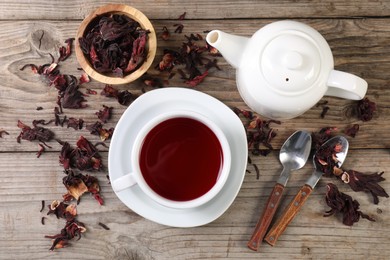 Photo of Delicious hibiscus tea in cup, teapot, dry roselle sepals and spoons on wooden table, flat lay