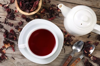 Photo of Delicious hibiscus tea in cup, teapot, dry roselle sepals and spoons on wooden table, flat lay