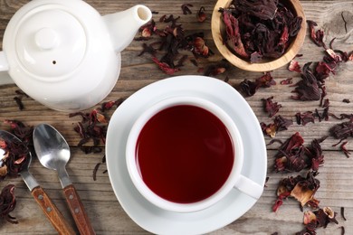 Photo of Delicious hibiscus tea in cup, teapot, dry roselle sepals and spoons on wooden table, flat lay
