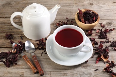 Photo of Delicious hibiscus tea in cup, teapot, dry roselle sepals and spoons on wooden table