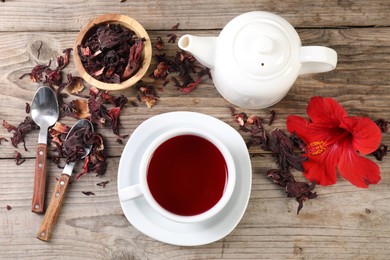 Photo of Delicious hibiscus tea in cup, teapot, dry roselle sepals and flower on wooden table, flat lay