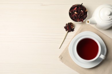 Photo of Delicious hibiscus tea in cup, teapot, dry roselle sepals and spoon on light wooden table, flat lay. Space for text