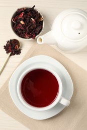 Photo of Delicious hibiscus tea in cup, teapot, dry roselle sepals and spoon on light wooden table, flat lay