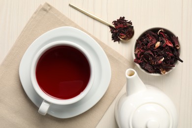 Photo of Delicious hibiscus tea in cup, teapot, dry roselle sepals and spoon on light wooden table, flat lay