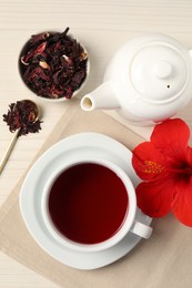 Photo of Delicious hibiscus tea in cup, teapot, dry roselle sepals and flower on light wooden table, flat lay