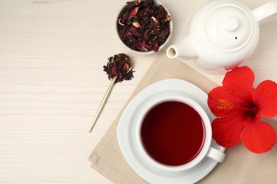 Photo of Delicious hibiscus tea in cup, teapot, dry roselle sepals and flower on light wooden table, flat lay. Space for text