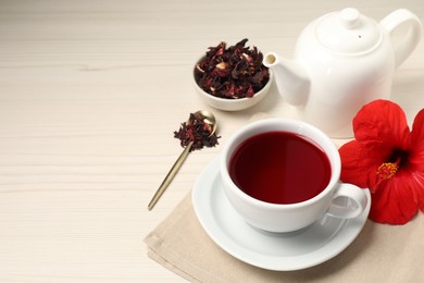 Photo of Delicious hibiscus tea in cup, teapot, dry roselle sepals and flower on light wooden table, space for text