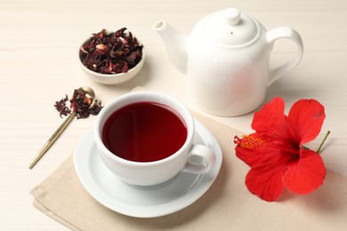 Photo of Delicious hibiscus tea in cup, teapot, dry roselle sepals and flower on light wooden table