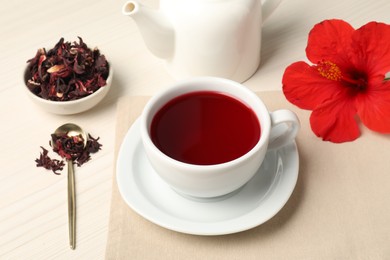 Photo of Delicious hibiscus tea in cup, teapot, dry roselle sepals and flower on light wooden table, closeup