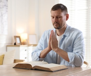 Photo of Man with Bible praying at table indoors. Space for text