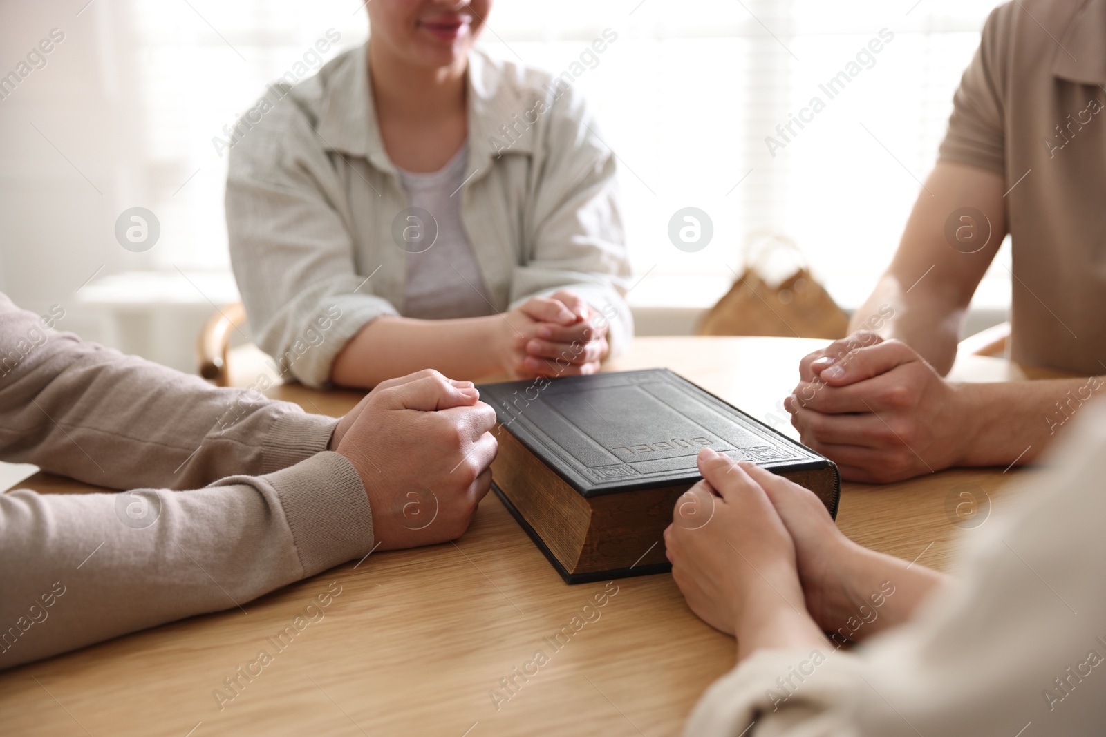 Family with Bible praying at wooden table indoors, closeup Photo of Family with Bible praying at wooden table indoors, closeup