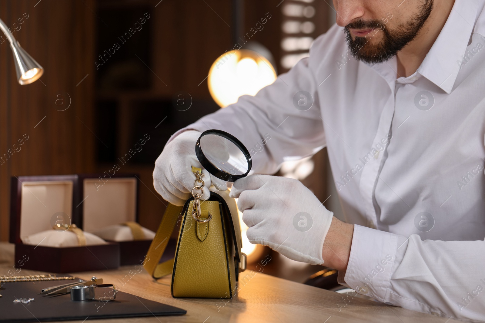 Appraiser with magnifying glass evaluating leather bag at table indoors, closeup Photo of Appraiser with magnifying glass evaluating leather bag at table indoors, closeup