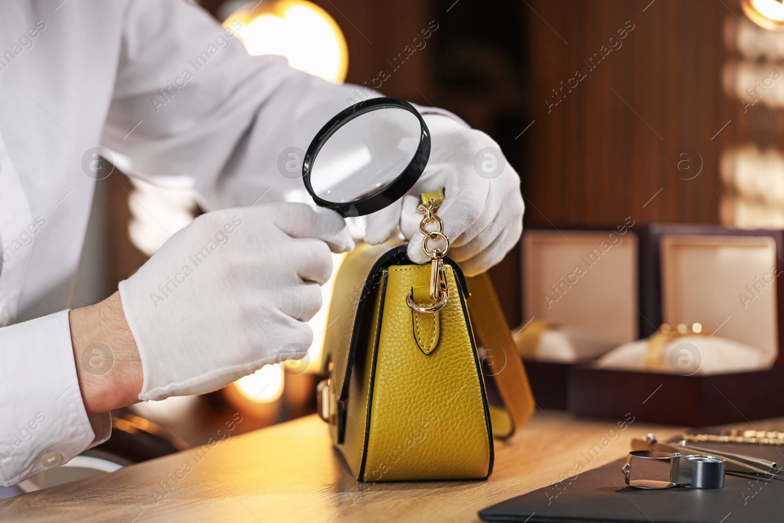 Appraiser with magnifying glass evaluating leather bag at table indoors, closeup Photo of Appraiser with magnifying glass evaluating leather bag at table indoors, closeup