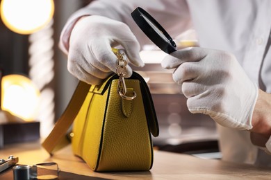 Photo of Appraiser with magnifying glass evaluating leather bag at table indoors, closeup