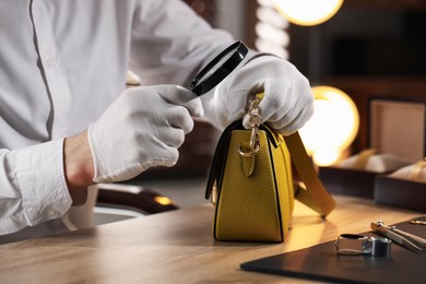 Photo of Appraiser with magnifying glass evaluating leather bag at table indoors, closeup