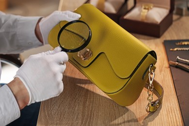 Photo of Appraiser with magnifying glass evaluating leather bag at table indoors, closeup