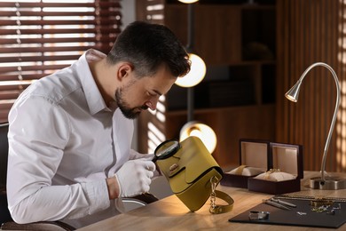 Photo of Appraiser with magnifying glass evaluating leather bag at table indoors