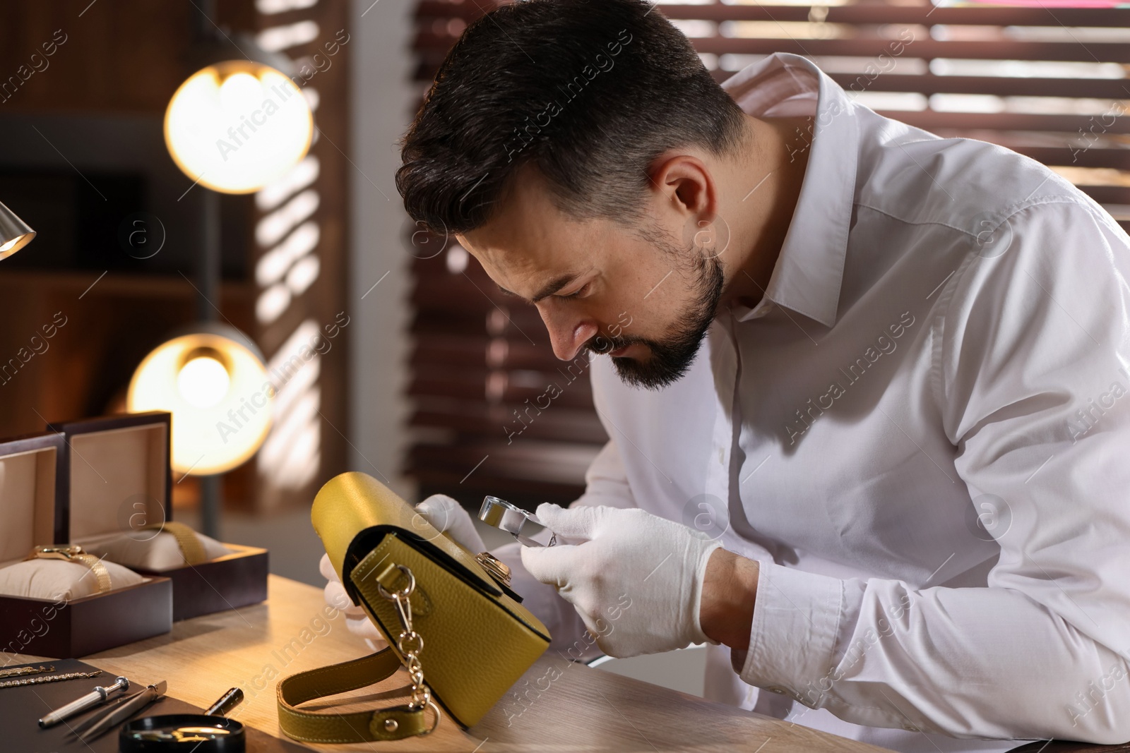 Appraiser with loupe evaluating leather bag at table indoors Photo of Appraiser with loupe evaluating leather bag at table indoors