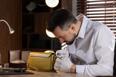 Photo of Appraiser with loupe evaluating leather bag at table indoors