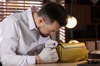 Photo of Appraiser with loupe evaluating leather bag at table indoors