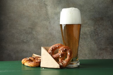 Photo of Tasty pretzels and glass of beer on green wooden table against grey textured background, closeup