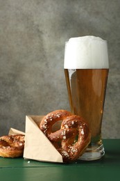 Photo of Tasty pretzels and glass of beer on green wooden table against grey textured background, closeup