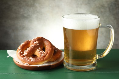 Photo of Tasty pretzels and glass of beer on green wooden table against grey textured background, closeup