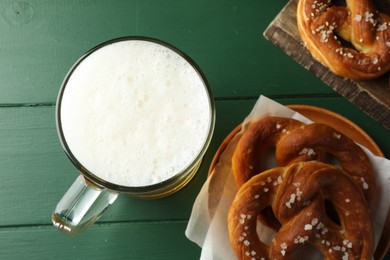Photo of Tasty pretzels and glass of beer on green wooden table, flat lay