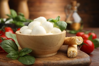 Photo of Tasty mozzarella cheese balls, breadsticks, tomatoes and basil on wooden table, closeup