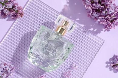 Photo of Bottle of perfume and beautiful lilac flowers on light violet background, flat lay