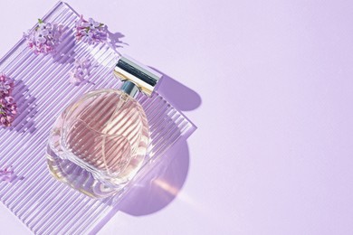 Photo of Bottle of perfume and beautiful lilac flowers on light violet background, flat lay. Space for text