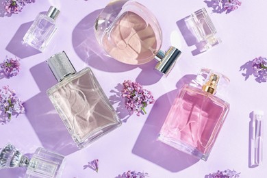 Photo of Luxury perfumes and beautiful lilac flowers on light violet background, flat lay