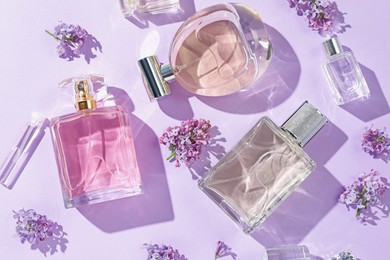 Photo of Luxury perfumes and beautiful lilac flowers on light violet background, flat lay