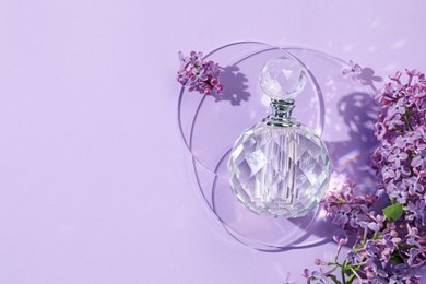 Photo of Bottle of perfume and beautiful lilac flowers on light violet background, flat lay. Space for text