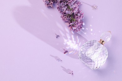 Photo of Bottle of perfume and beautiful lilac flowers on light violet background, flat lay. Space for text