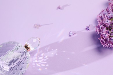Photo of Bottle of perfume and beautiful lilac flowers on light violet background, closeup
