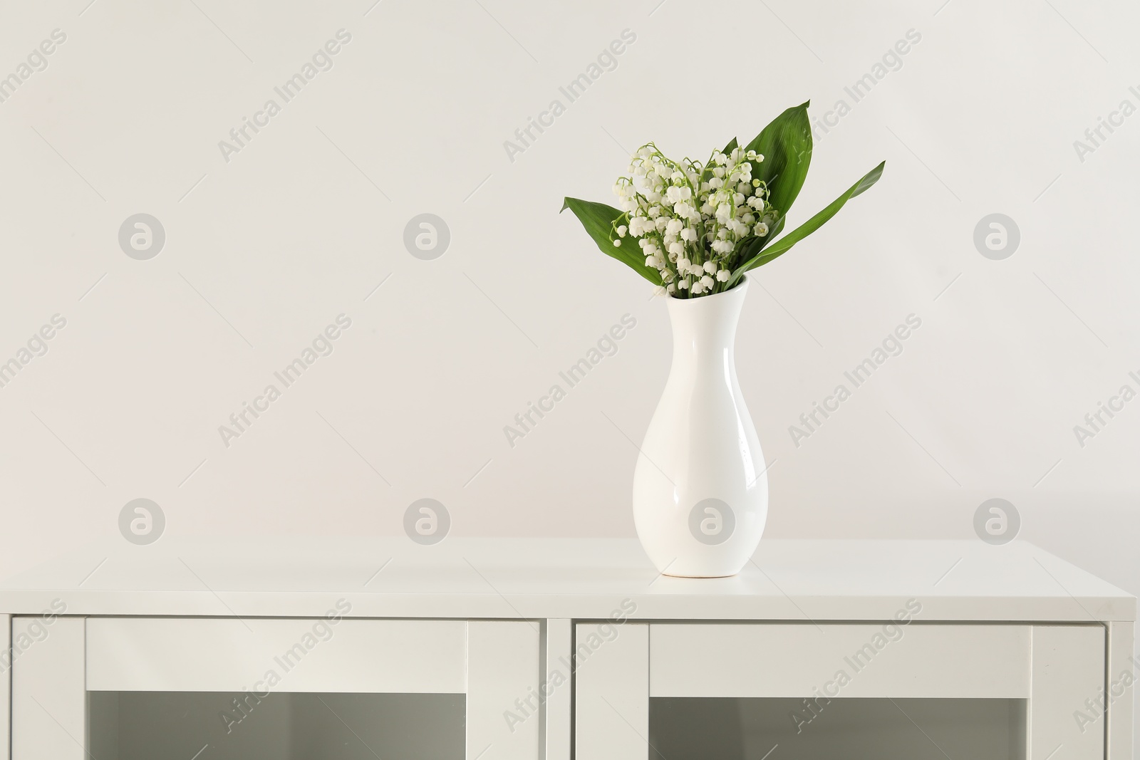 Beautiful lilies of the valley flowers in vase on cabinet. Space for text Photo of Beautiful lilies of the valley flowers in vase on cabinet. Space for text