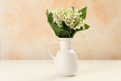Photo of Beautiful lilies of the valley flowers in vase on white table near beige wall