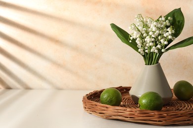 Photo of Beautiful lilies of the valley flowers in vase and limes on white table near beige wall. Space for text